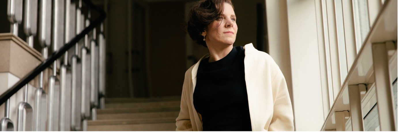 Editorial portrait of Teresa Zimmermann in a cream coat standing in a bright columned interior, looking toward the light