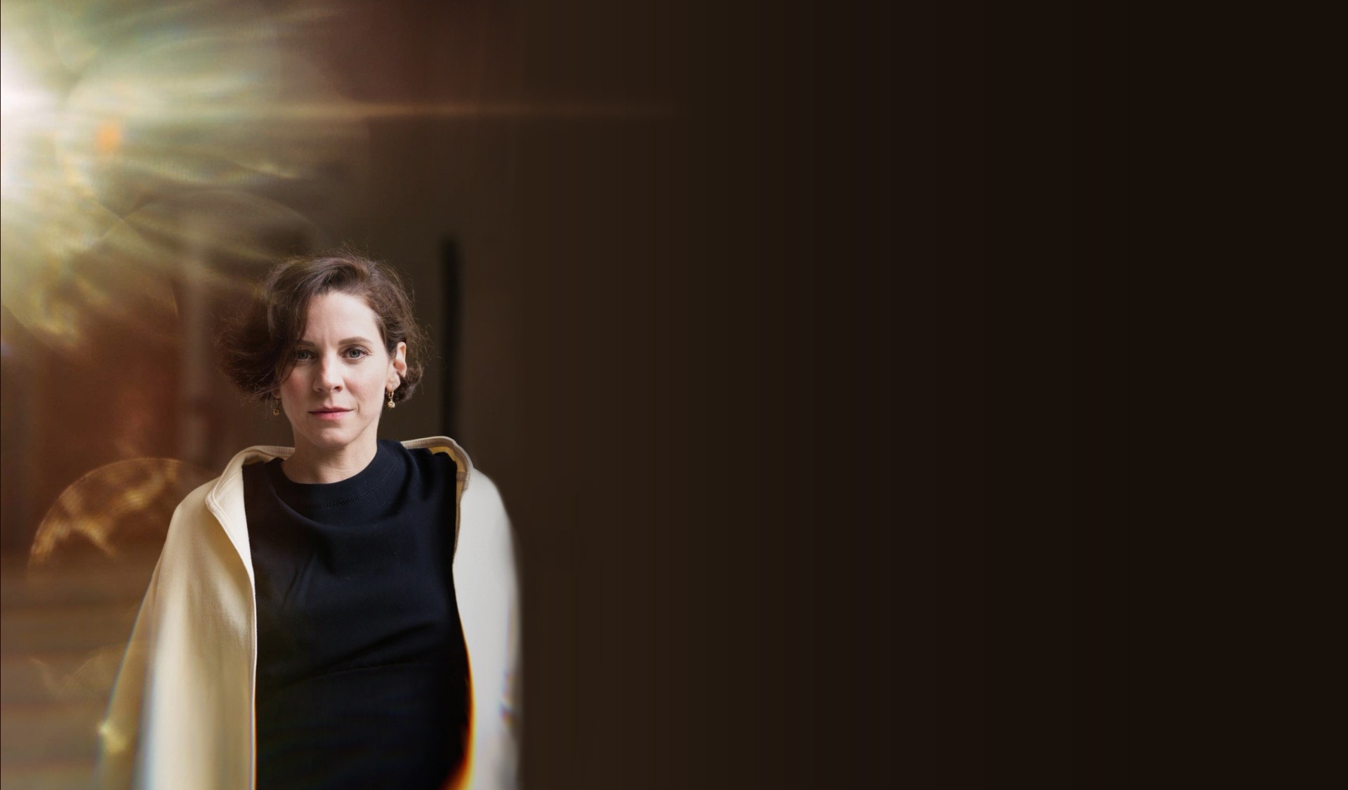 Editorial portrait of Teresa Zimmermann in a cream coat with warm light entering from the left, standing in a dark interior